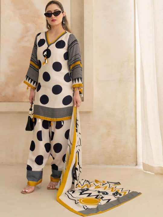 A woman modeling a three-piece traditional outfit with a cream base, black polka dots, gray stripes, and mustard yellow accents, including a matching dupatta, in a warm beige interior setting.