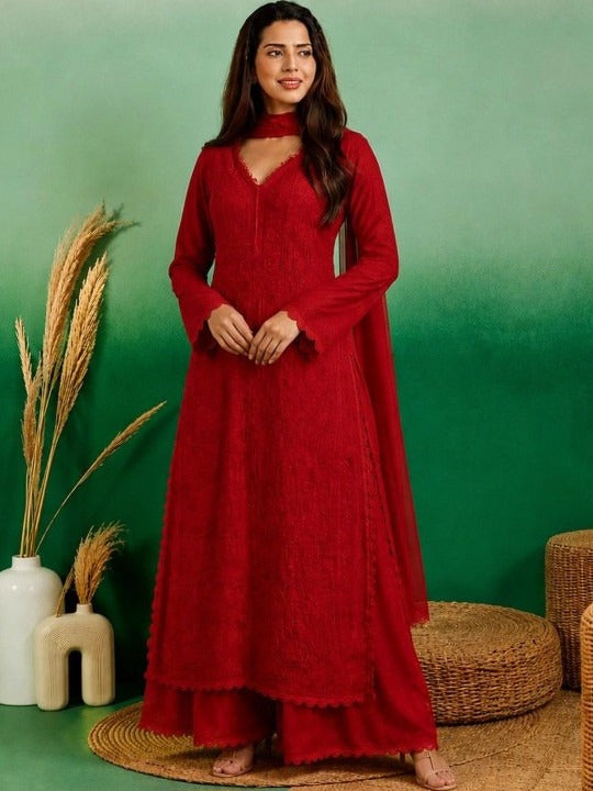 A woman stands barefoot against a green backdrop wearing a long-sleeve red maxi dress with a high neck and lace-like texture. Decorative dried grass in white vases and woven round cushions appear at the sides.